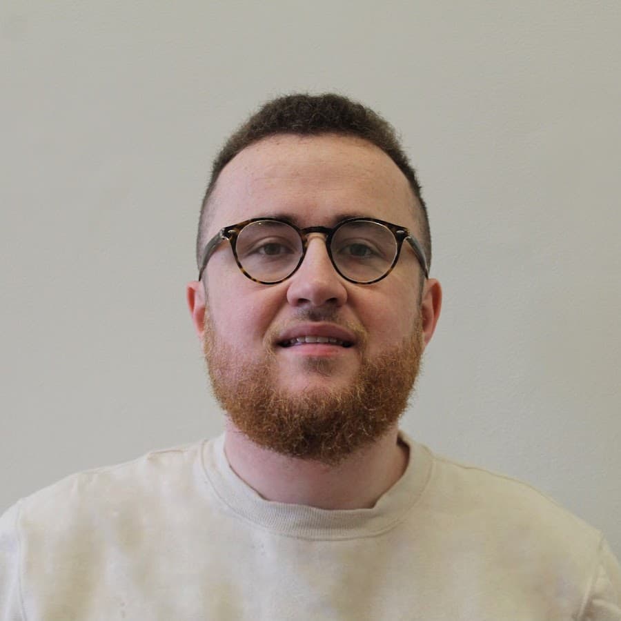 A man (Robert James Gabriel) with short-cropped hair and a beard, wearing round glasses and a light-colored shirt, smiles slightly against a plain white background.